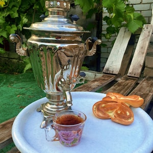 May include: A polished silver samovar with decorative elements, a glass teacup with a floral pattern, and two golden pastries are on a white tray. The tray is on a wooden surface, with green leaves in the background.