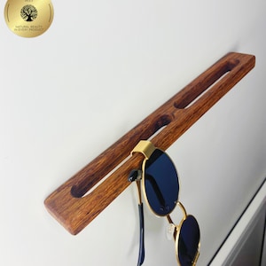 May include: A wooden sunglasses holder with a pair of sunglasses hanging from it. The holder is a rich brown colour and has two slots carved into it. The sunglasses have gold frames and dark blue lenses. The holder is mounted on a white wall.