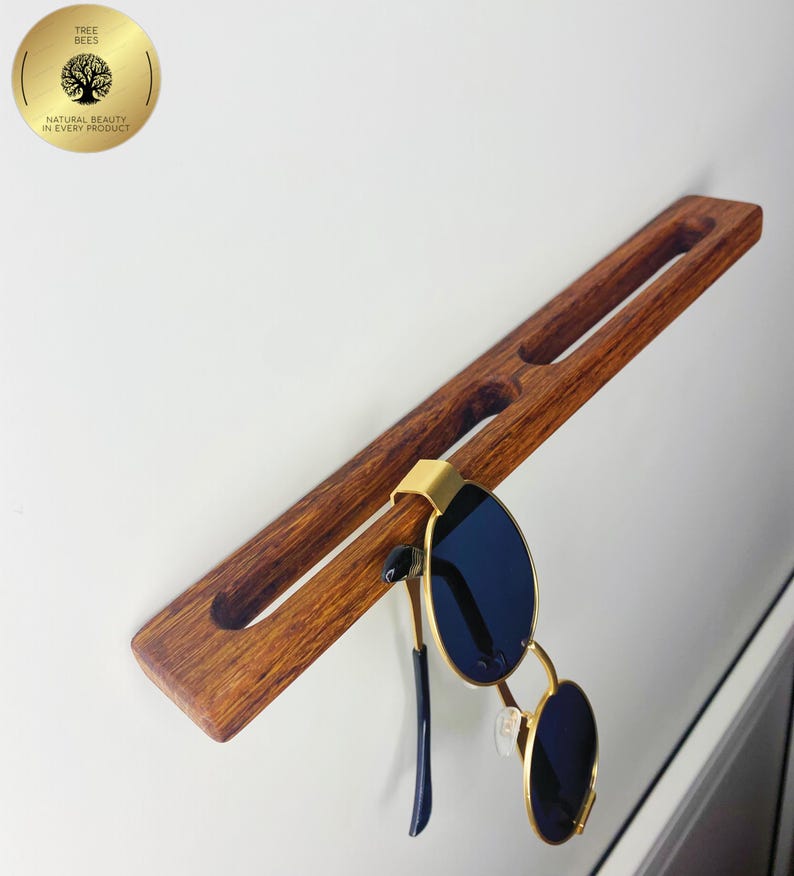 May include: A wooden sunglasses holder with a pair of sunglasses hanging from it. The holder is a rich brown colour and has two slots carved into it. The sunglasses have gold frames and dark blue lenses. The holder is mounted on a white wall.