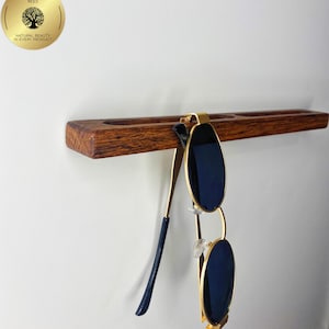 May include: A wooden sunglasses holder with a pair of sunglasses hanging from it. The holder is made of dark brown wood and has a gold-coloured metal clip. The sunglasses have gold frames and dark blue lenses. The holder is mounted on a white wall.