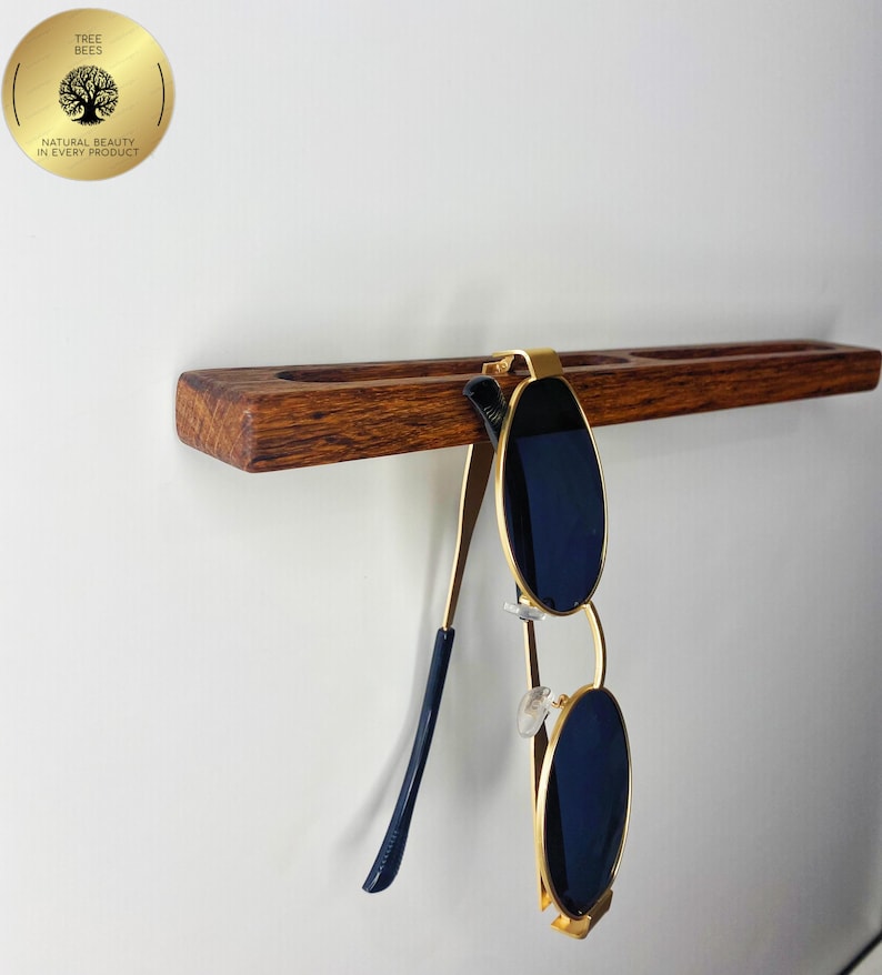 May include: A wooden sunglasses holder with a pair of sunglasses hanging from it. The holder is made of dark brown wood and has a gold-coloured metal clip. The sunglasses have gold frames and dark blue lenses. The holder is mounted on a white wall.