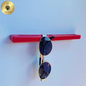May include: A red sunglasses holder with a pair of sunglasses hanging from it. The sunglasses have gold frames and dark lenses. The holder is mounted on a white wall. A gold sticker with the text "Tree Bees" is in the upper left corner.