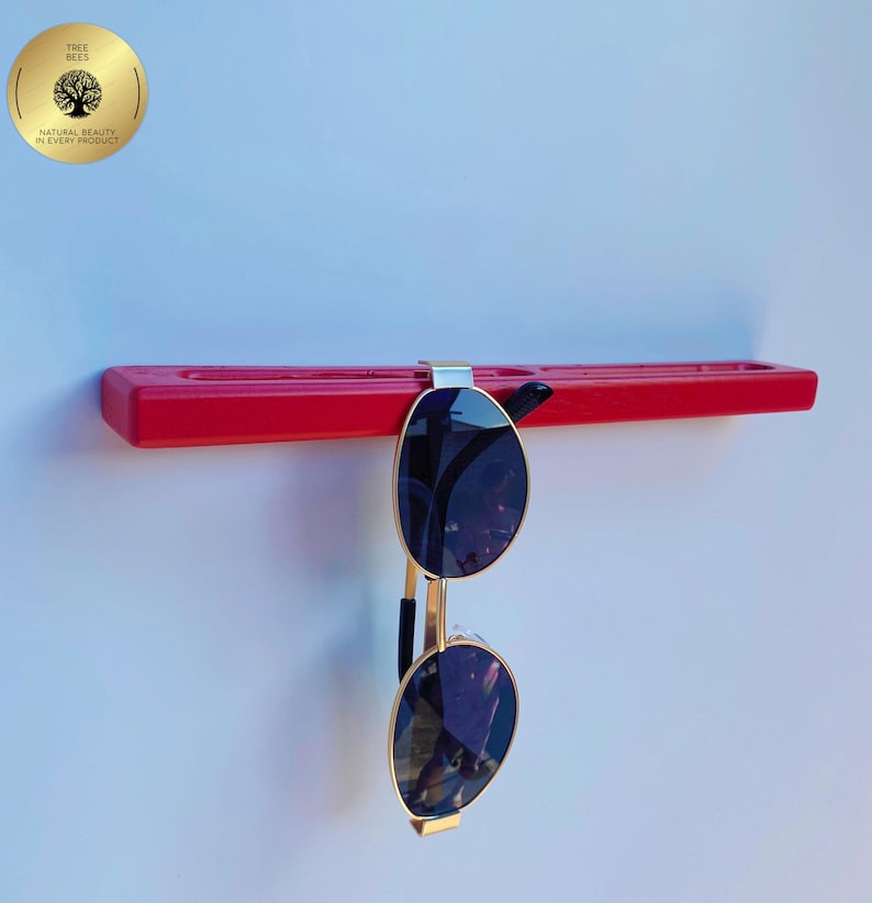 May include: A red sunglasses holder with a pair of sunglasses hanging from it. The sunglasses have gold frames and dark lenses. The holder is mounted on a white wall. A gold sticker with the text "Tree Bees" is in the upper left corner.