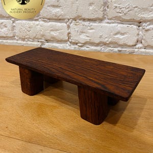May include: A dark brown wooden platform with four short, rectangular legs. The platform has a smooth, rectangular top and is made of a wood with a visible grain. The platform is displayed on a light brown wooden surface.