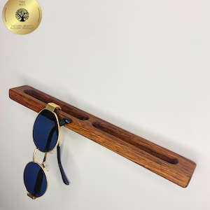 May include: A wooden sunglasses holder with three circular indentations and a pair of blue-lensed sunglasses with gold frames. The holder is made of dark brown wood and is mounted on a white wall. A gold circular logo is in the upper left corner.