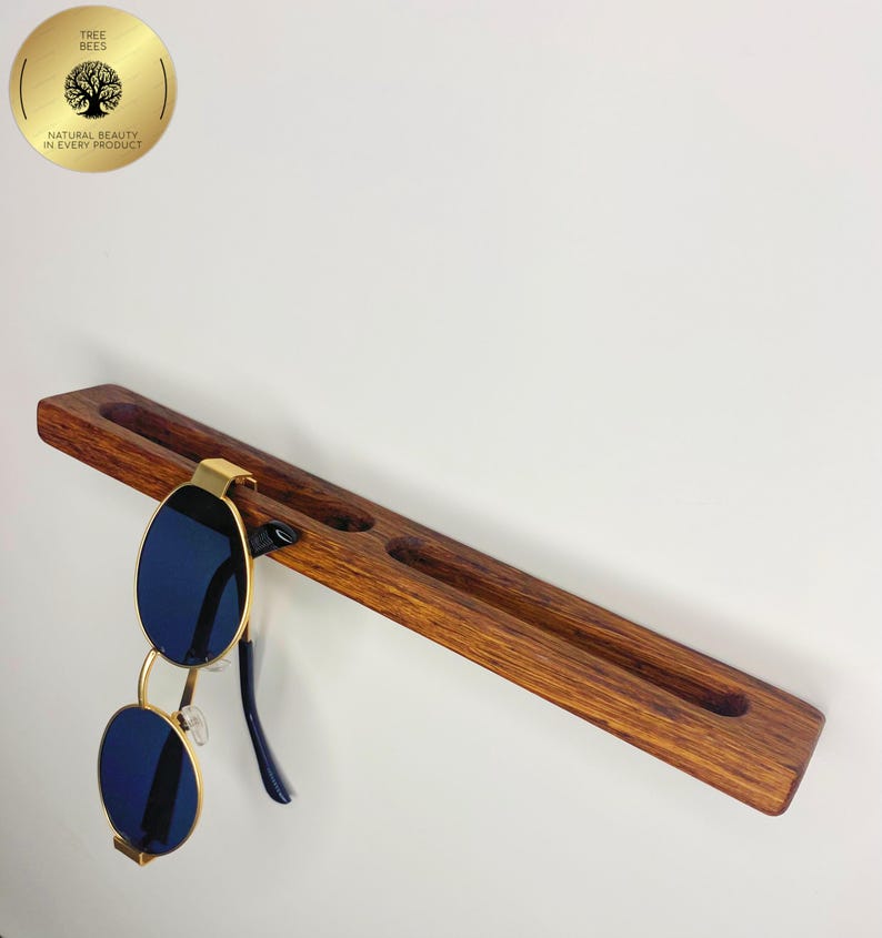 May include: A wooden sunglasses holder with three circular indentations and a pair of blue-lensed sunglasses with gold frames. The holder is made of dark brown wood and is mounted on a white wall. A gold circular logo is in the upper left corner.