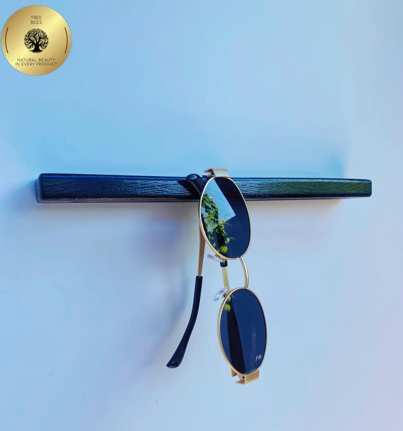 May include: A black wooden shelf with gold-framed sunglasses hanging from it. The sunglasses have dark lenses. A gold circular logo with the text "TREE BEES" and "NATURAL BEAUTY IN EVERY PRODUCT" is in the upper left corner.