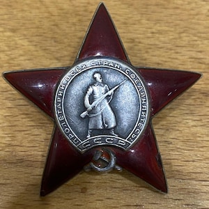 May include: A red enameled star-shaped medal with a central silver medallion depicting a soldier with a rifle. The medallion is surrounded by text in a circular pattern. A hammer and sickle emblem is at the bottom.