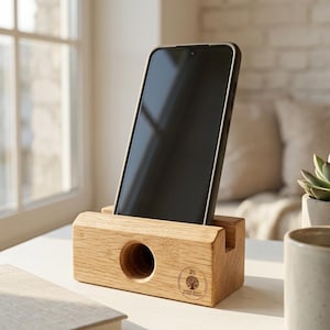 Puede incluir: Un soporte de teléfono de madera de color marrón claro rectangular con una ranura horizontal para un teléfono inteligente y un orificio circular amplificador de sonido en la parte frontal. Un logotipo circular con un árbol y el texto Tree Beer Natural Beauty está impreso.