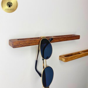 May include: A wooden sunglasses holder with a pair of sunglasses hanging from it. The holder is a dark brown colour and has a groove for the sunglasses to rest in. The sunglasses have gold frames and dark blue lenses.