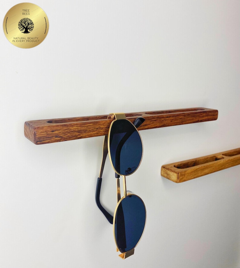May include: A wooden sunglasses holder with a pair of sunglasses hanging from it. The holder is a dark brown colour and has a groove for the sunglasses to rest in. The sunglasses have gold frames and dark blue lenses.