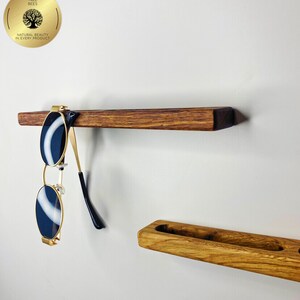 May include: A wooden shelf with sunglasses hanging from it. The sunglasses have a gold frame and dark blue lenses. The shelf is made of dark brown wood. A second wooden shelf is below the first shelf. The image includes a gold emblem with the text "TREE BEES NATURAL BEAUTY IN EVERY PRODUCT."