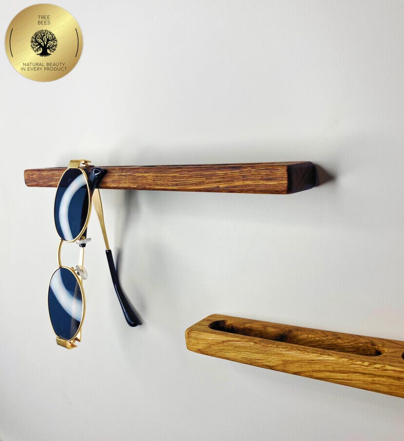 May include: A wooden shelf with sunglasses hanging from it. The sunglasses have a gold frame and dark blue lenses. The shelf is made of dark brown wood. A second wooden shelf is below the first shelf. The image includes a gold emblem with the text "TREE BEES NATURAL BEAUTY IN EVERY PRODUCT."