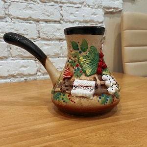 Op de afbeelding: Een decoratieve keramische koffiepot met een lange zwarte handgreep. De pot is beige met kleurrijke geschilderde details, waaronder rode bessen, groene bladeren en witte bloemaccenten. De pot staat op een houten tafel.