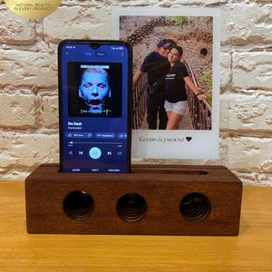 May include: A dark brown wooden phone stand with three circular speaker holes. A smartphone displays a music app with the song "Du Hast" by Rammstein. A photo of a couple is behind the phone.