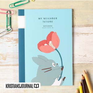 May include: A light blue notebook with the text "MY NEIGHBOR TOTORO" and a graphic of Totoro holding a flower. The notebook has a blue spine and the text "NOTEBOOK GRAPH PAPER B6". The bottom left corner has the text "KRISTIANSJOURNAL".