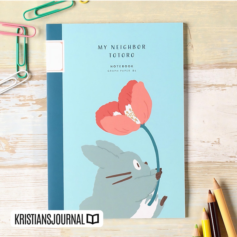 May include: A light blue notebook with the text "MY NEIGHBOR TOTORO" and a graphic of Totoro holding a flower. The notebook has a blue spine and the text "NOTEBOOK GRAPH PAPER B6". The bottom left corner has the text "KRISTIANSJOURNAL".