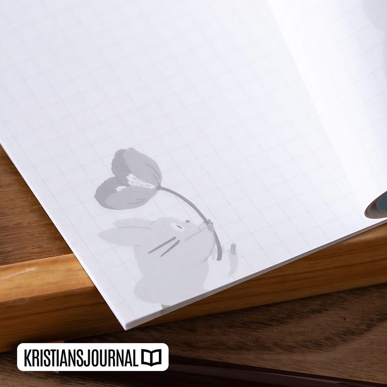 May include: Open notebook with a grid pattern. A gray illustration of a rabbit holding a flower is in the corner. The text "KRISTIANSJOURNAL" is at the bottom. The notebook is open on a wooden surface.