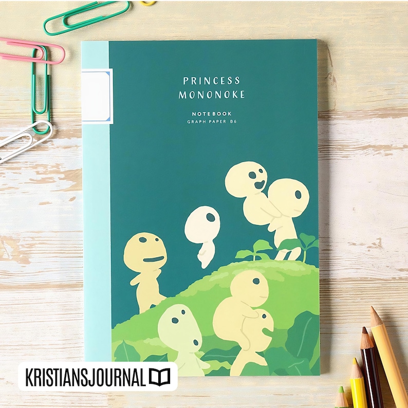 May include: A notebook with a teal cover featuring the text "PRINCESS MONONOKE NOTEBOOK GRAPH PAPER 06." The cover has an illustration of several light yellow spirit-like figures on a green hill. The notebook is on a light wood surface with paperclips and pencils.