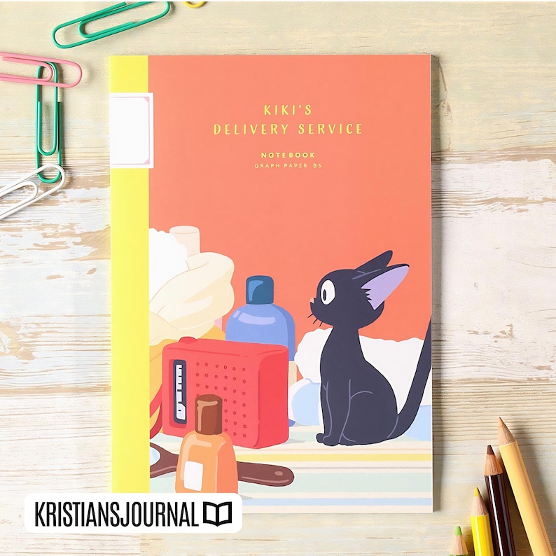 May include: A notebook with a yellow spine and an orange cover featuring the text "Kiki's Delivery Service." The cover also displays a black cat, a red radio, and other illustrations. The notebook is labeled "Notebook Graph Paper 80."