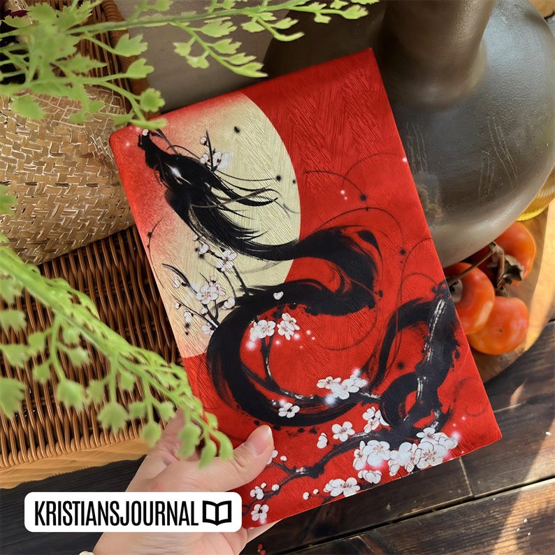 K&ouml;nnte beinhalten: Ein rotes Notizbuch mit einer schwarz-wei&szlig;en Illustration eines Drachen und Kirschbl&uuml;ten. Das Notizbuch hat einen goldenen Mondhintergrund und den Text "KRISTIANSJOURNAL".