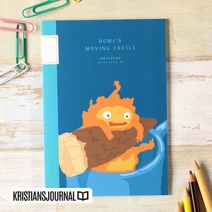 May include: A blue notebook with the text "Howl's Moving Castle" and a cartoon illustration of Calcifer. The notebook has a light blue spine and a white label. The words "Notebook" and "Graph Paper 80" are also printed on the cover.