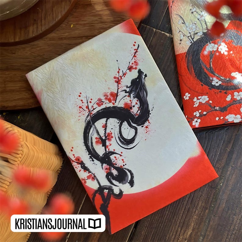 K&ouml;nnte beinhalten: Ein Journal mit einer Drachenillustration und dem Text "KRISTIANSJOURNAL". Das Cover zeigt ein schwarzes Drachendesign mit roten Akzenten und Kirschbl&uuml;ten auf wei&szlig;em und rotem Hintergrund. Das Journal liegt auf einer Holzoberfl&auml;che.