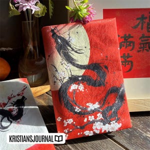 K&ouml;nnte beinhalten: Rotes Notizbuch mit Drachen- und Monddesign. Das Notizbuch zeigt einen schwarzen Drachen mit wei&szlig;en Kirschbl&uuml;ten vor rotem Hintergrund. Der Text "KRISTIANSJOURNAL" ist unten sichtbar. Eine Vase mit Blumen und ein Kunstwerk mit chinesischen Schriftzeichen befinden sich im Hintergrund.