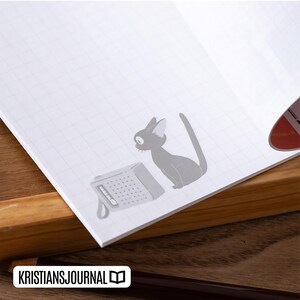 May include: Open notebook with a grid pattern, featuring a gray cat illustration and a small radio graphic. The notebook is on a wooden surface, with the text "KRISTIANSJOURNAL" at the bottom.