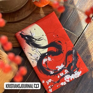 K&ouml;nnte beinhalten: Rotes Notizbuch mit einer schwarzen Drachenillustration, wei&szlig;en Kirschbl&uuml;ten und einem gelben Monddesign. Das Notizbuch hat den Text "KRISTIANSJOURNAL" in einem wei&szlig;en Rechteck unten. Auf einer Holzoberfl&auml;che.