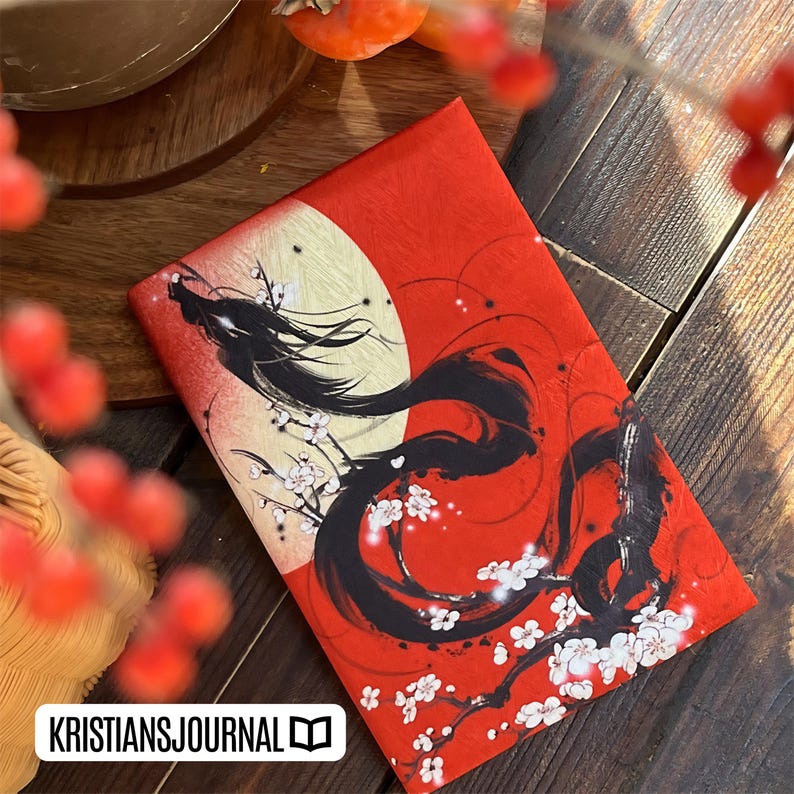 K&ouml;nnte beinhalten: Rotes Notizbuch mit einer schwarzen Drachenillustration, wei&szlig;en Kirschbl&uuml;ten und einem gelben Monddesign. Das Notizbuch hat den Text "KRISTIANSJOURNAL" in einem wei&szlig;en Rechteck unten. Auf einer Holzoberfl&auml;che.