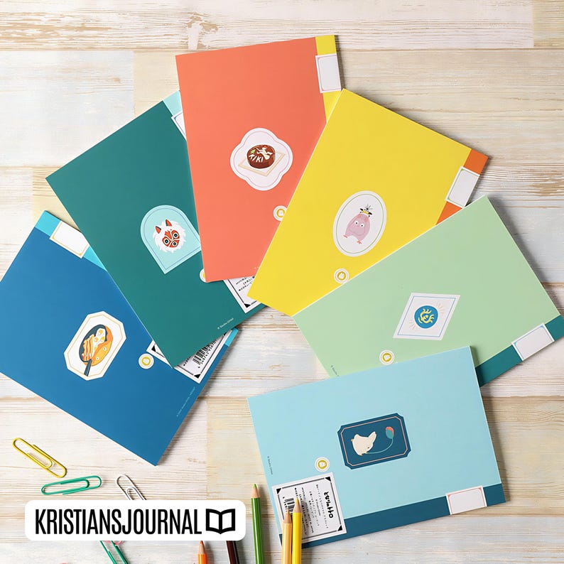 May include: A collection of colorful notebooks in various shades of blue, green, orange, and yellow. Each notebook features a unique sticker with illustrations. The notebooks are arranged on a light wooden surface, with pencils and paperclips nearby.