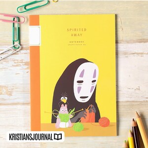 May include: A yellow notebook with the words "Spirited Away" and "Notebook Graph Paper B6" printed on the cover. The cover features an illustration of a character from the movie, with a black cloak and white mask, knitting with yarn.