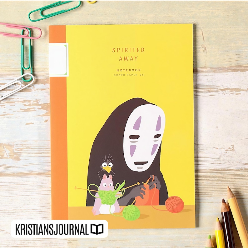 May include: A yellow notebook with the words "Spirited Away" and "Notebook Graph Paper B6" printed on the cover. The cover features an illustration of a character from the movie, with a black cloak and white mask, knitting with yarn.