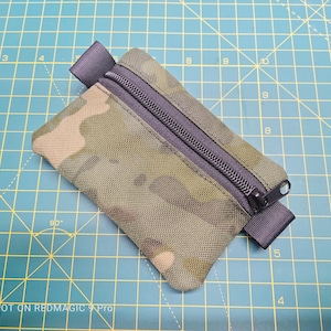 Peut inclure: Une petite pochette zippée de camouflage vert et marron avec une fermeture éclair noire et une sangle noire. La pochette est posée sur une surface quadrillée verte.