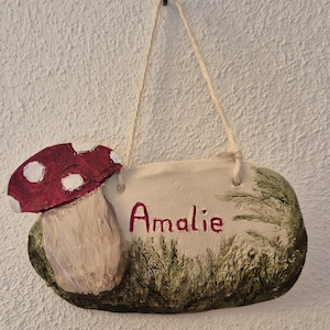 Può includere: Un cartello in argilla dipinto a mano con un motivo a fungo e il nome "Amalie" scritto in vernice rossa. Il cartello è appeso a una corda e ha uno sfondo verde e marrone.
