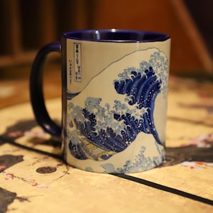 May include: Ceramic mug featuring a blue and white wave design, with a dark blue handle and interior. The mug's design is inspired by Japanese art, with a detailed depiction of a large wave. The mug sits on a gold-colored surface.