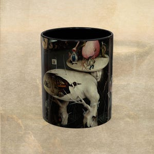 May include: Black ceramic coffee mug featuring a surrealist painting by Hieronymus Bosch. The artwork depicts a white creature with a broken shell, figures, and a circular platform with more figures. The mug has a black interior.