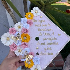 May include: A light pink graduation cap decorated with paper flowers in white, yellow, pink, and blue. Gold butterflies and text in gold script are also featured. The text reads "Un logro mas con la bendicion de Dios, el sacrificio de mi familia y mi dedicacion."