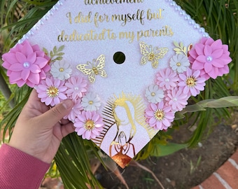 May include: A decorated graduation cap with the text "This is a achievement I did for myself but dedicated to my parents". The cap is light pink with gold accents, pink flowers, and gold butterfly decorations.