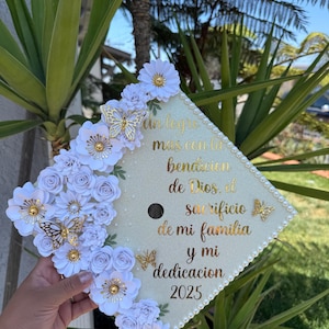 May include: A decorated graduation cap with white flowers, gold butterflies, and text in gold script. The cap is adorned with pearls around the edge and features the year 2025.