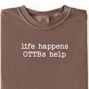 May include: A folded brown t-shirt with the text "life happens OTTBs help" in white. The shirt is made of a soft material and has a crew neck. The shirt is displayed on a white background.