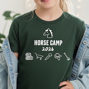 May include: A dark green t-shirt with the words "HORSE CAMP 2026" in white, along with horse-related icons. The shirt is worn by a young person, paired with a denim jacket.