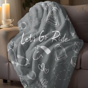 Puede incluir: Una manta de felpa gris con ilustraciones blancas de temática ecuestre, que incluyen herraduras y estribos. Las palabras "Let's Go Ride" están escritas en la manta con una fuente cursiva. La manta está sobre un sofá.