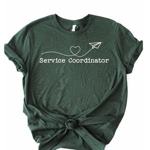 May include: A dark green t-shirt with a white graphic of a paper airplane with a dotted line trailing behind it, ending in a heart. The text "Service Coordinator" is printed below the graphic.