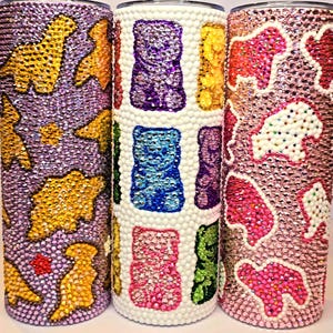 Może przedstawiać: Trzy kubki wysadzane kryształkami. Jeden jest fioletowy z żółtymi i pomarańczowymi kształtami dinozaurów. Drugi jest biały z kolorowymi kształtami żelków. Trzeci jest różowy z białymi i czerwonymi kształtami niedźwiedzi. Wszystkie są pokryte błyszczącymi kryształkami.