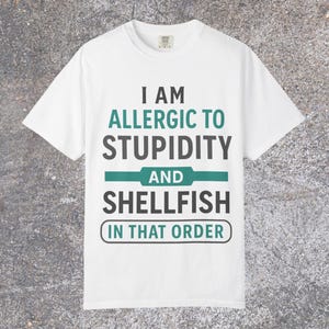 May include: White t-shirt with the text "I AM ALLERGIC TO STUPIDITY AND SHELLFISH IN THAT ORDER" in black and teal lettering. The shirt has a crew neck and short sleeves.