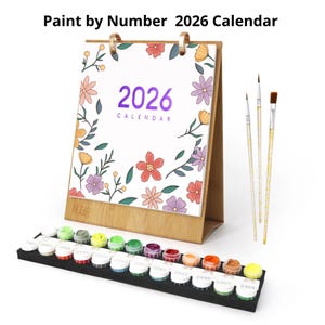 Puede incluir: Un calendario de pintar por números de 2026 con un diseño floral, acompañado de pinceles y botes de pintura numerados. El calendario muestra el año 2026 en morado, rodeado de flores coloridas. Los botes de pintura están dispuestos en un soporte negro.