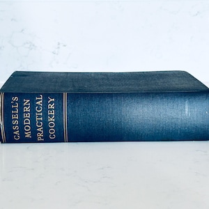 May include: A dark blue hardback book titled Cassell's Modern Practical Cookery. The book is positioned horizontally, displaying the spine with gold-coloured lettering against the dark blue fabric cover. The text is clearly visible on the spine.