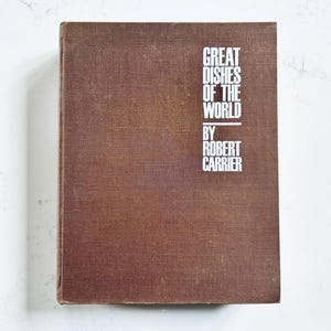 May include: A brown hardback book titled Great Dishes of the World by Robert Carrier. The front cover features a textured fabric finish with white sans-serif text printed in the upper right corner. The book is rectangular with a vertical orientation.
